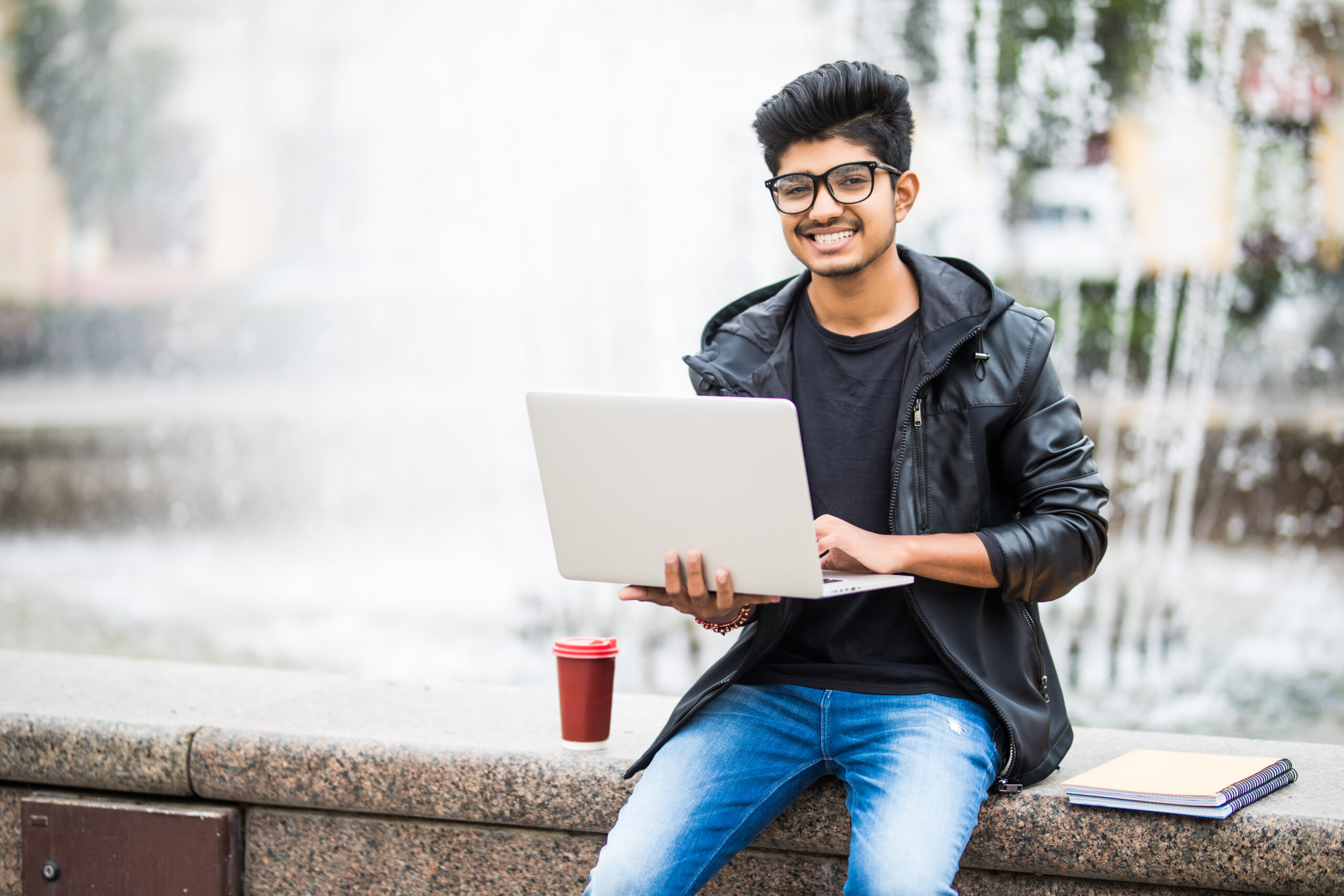 handsome indian man with laptop while sitting near the fountain in the city center on a day